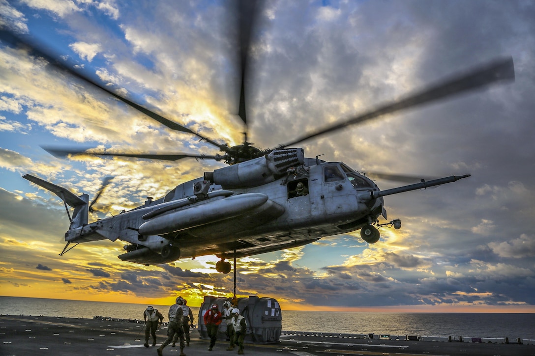 Service members on a ship's flight deck gather under a helicopter preparing to host an engine.