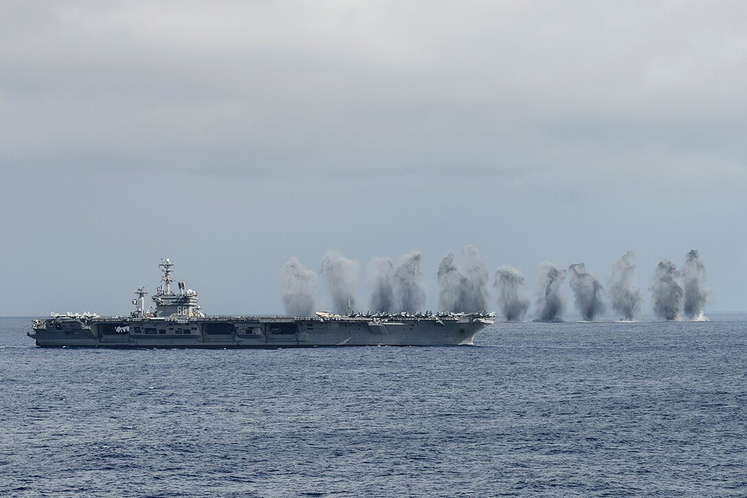 Smoke and water billow up from the ocean alongside an aircraft carrier.