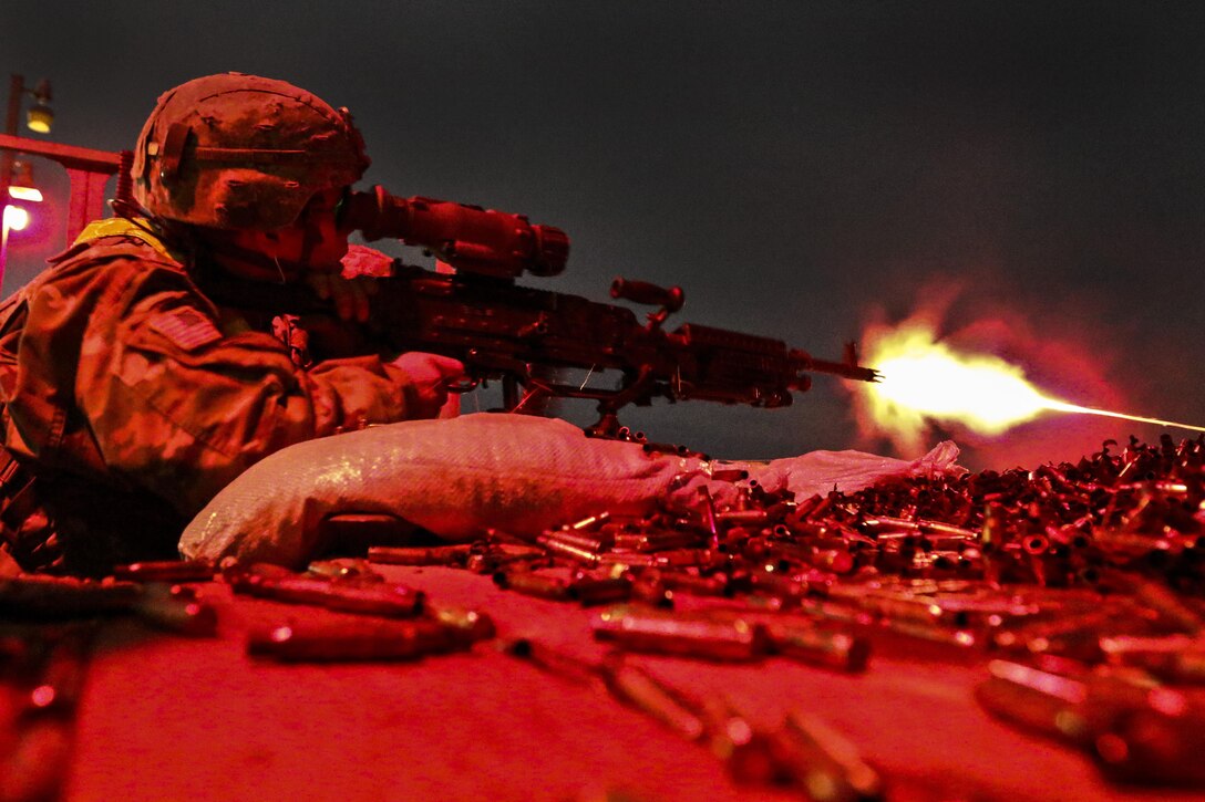 A soldier, illuminated by red light, fires a machine gun while prone on the ground in the dark.
