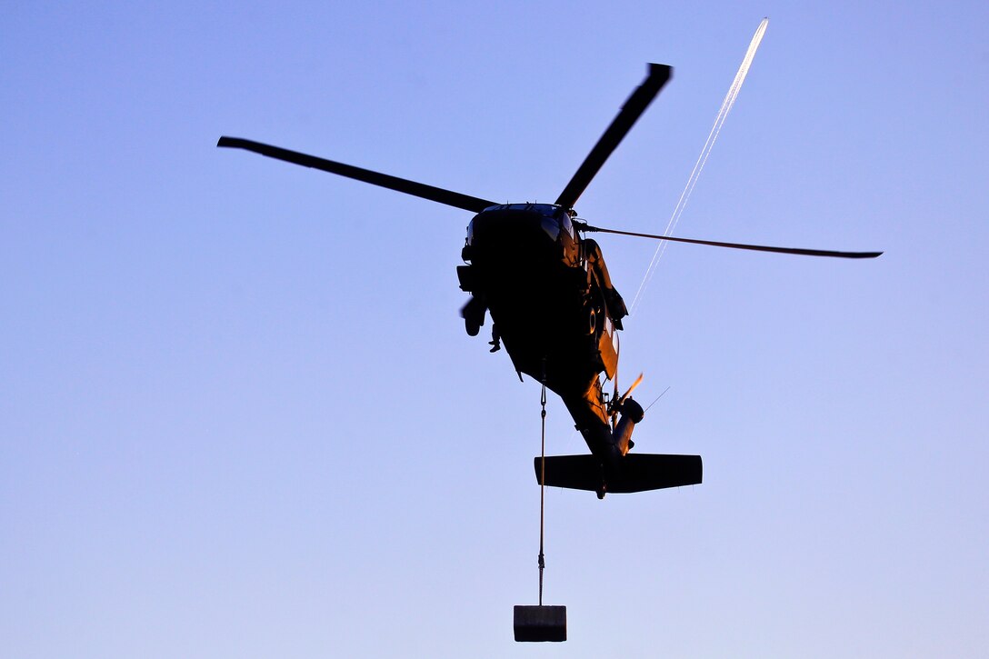 An Army UH-60 Black Hawk helicopter takes off from a clearing with a concrete block during slingload training exercise.