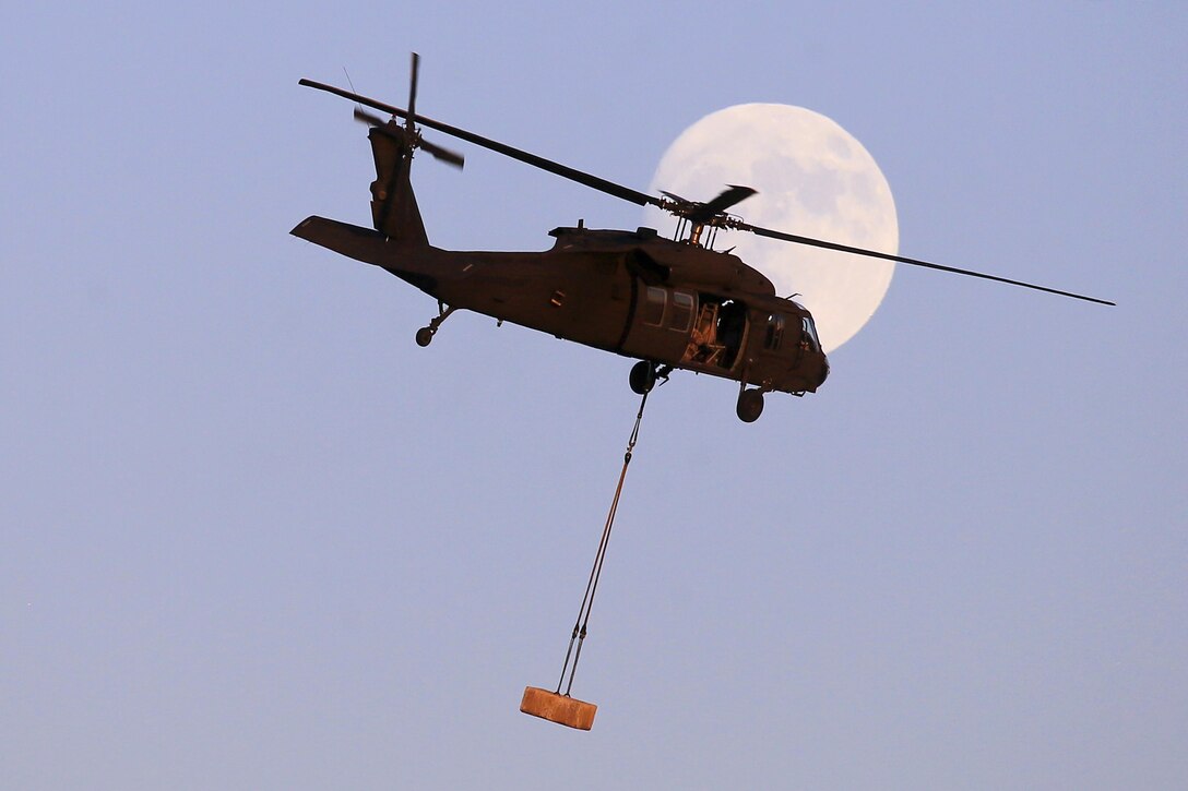 An Army UH-60 Black Hawk helicopter flies with a concrete block in front of the nearly full moon during slingload training exercise.