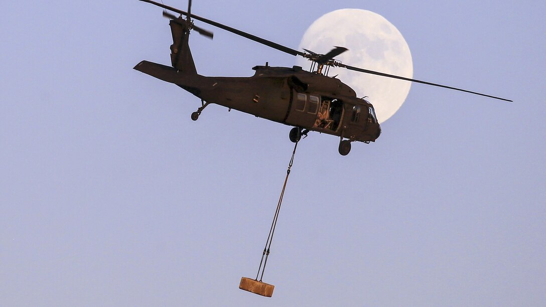 An Army helicopter flies with a concrete block in front of a nearly full moon during slingload training.
