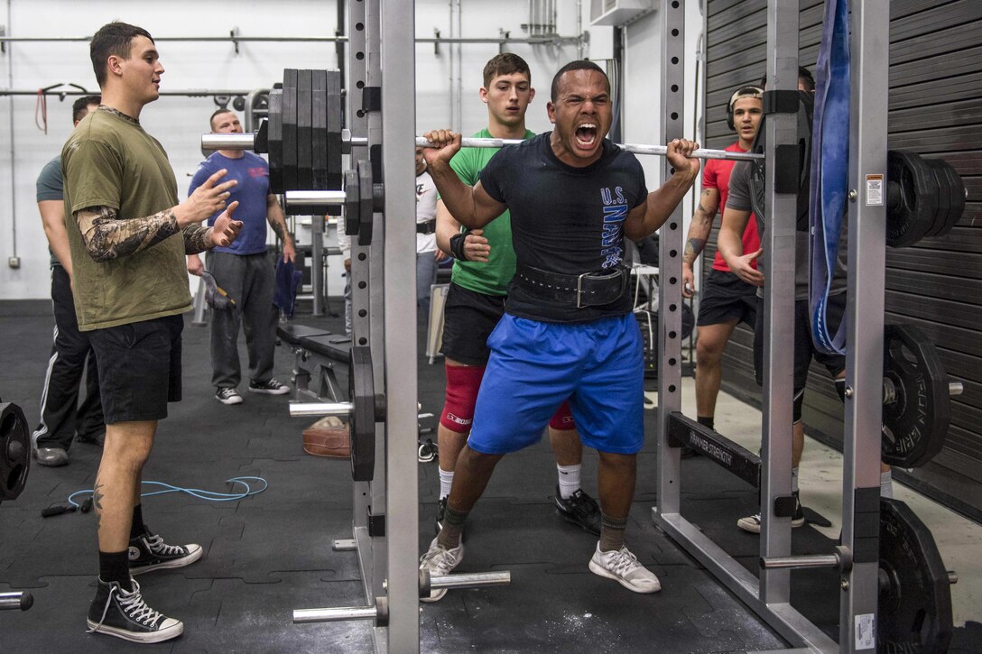 A sailor grimaces while lifting weights as others observe.