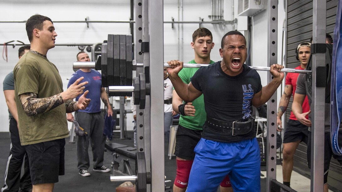 A sailor grimaces while lifting weights as others observe.
