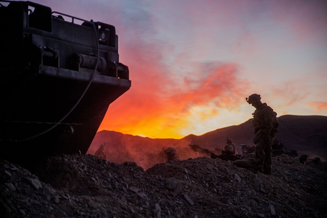 A Marine and a tactical vehicle, shown in silhouette against an orange-streaked sky, frame Marines digging a trench.