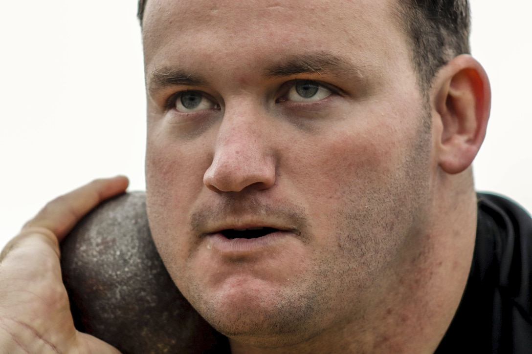 A soldier's face looks determined as he holds a shot put against it with one hand.