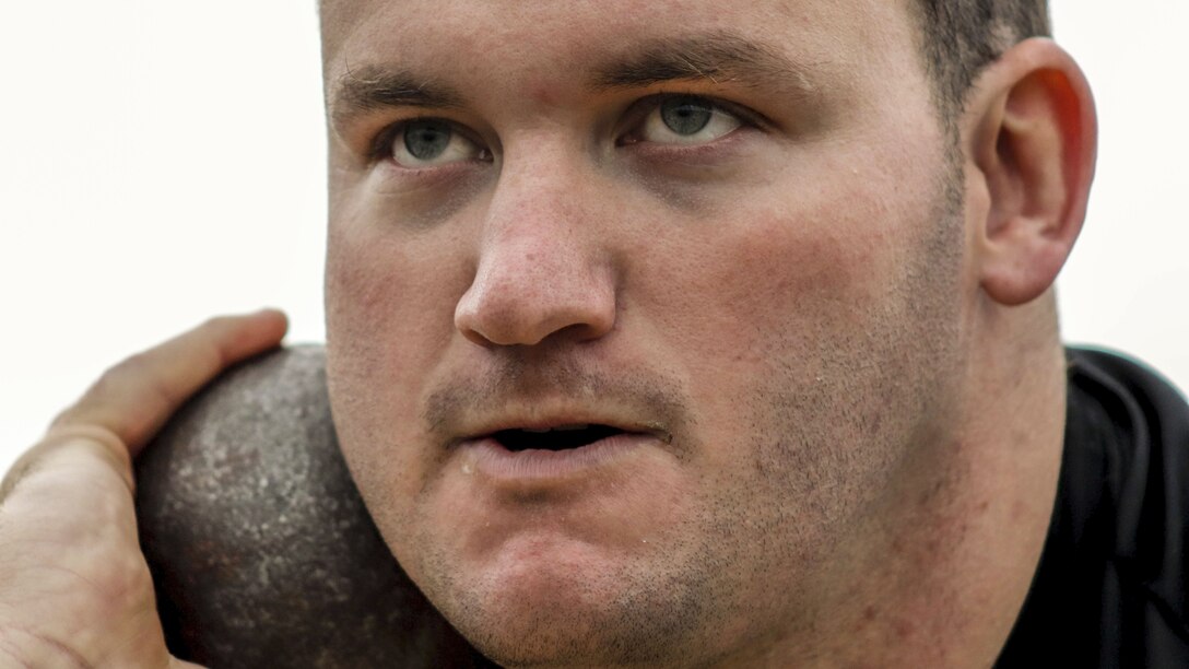 A soldier with a determined look on his face holds a shot put up against his neck with one hand.