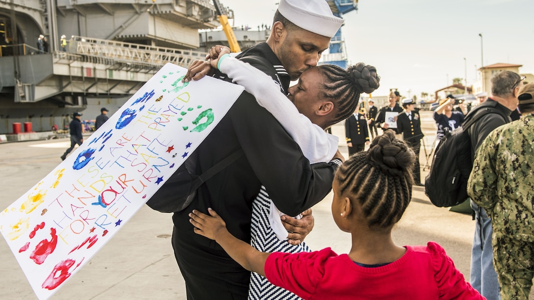 A sailor hugs his daughter while another runs toward him.