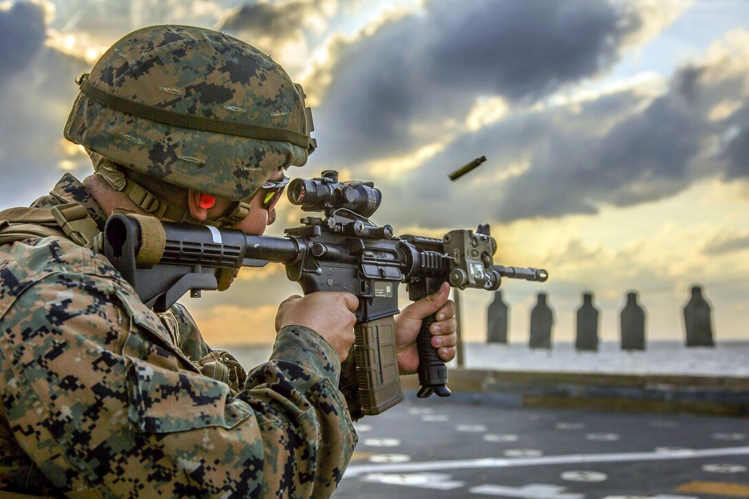 A Marine fires a carbine toward a hanging row of targets on a ship's flight deck.