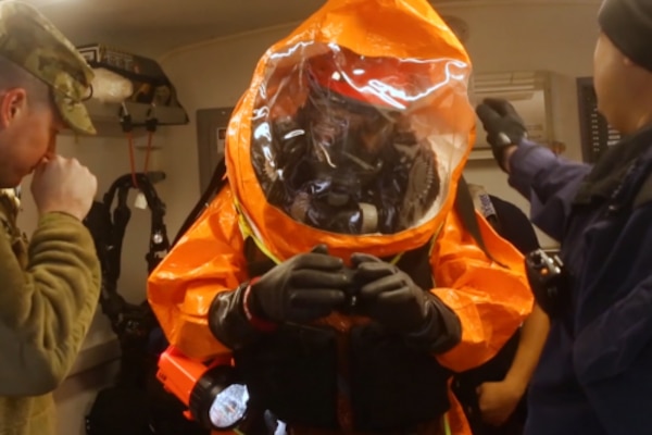 A service member wears a full-body orange protective suit and black gloves as two others flank him.