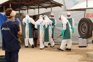 People in white, full-body protective gear walk with a woman into a makeshift entryway.
