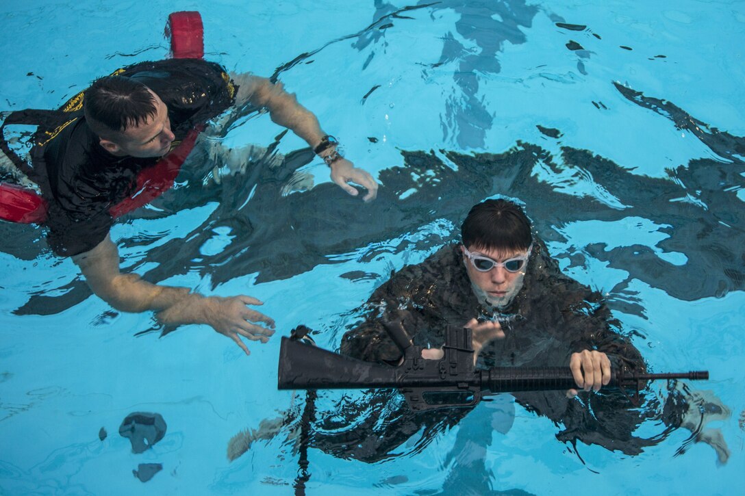 A Marine in a pool observes another Marine treading water while holding a rifle.