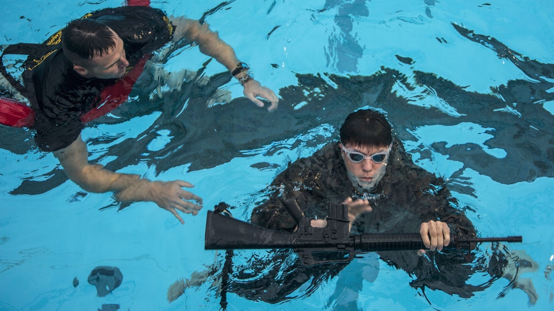 A Marine in a pool observes another Marine treading water while holding a rifle.