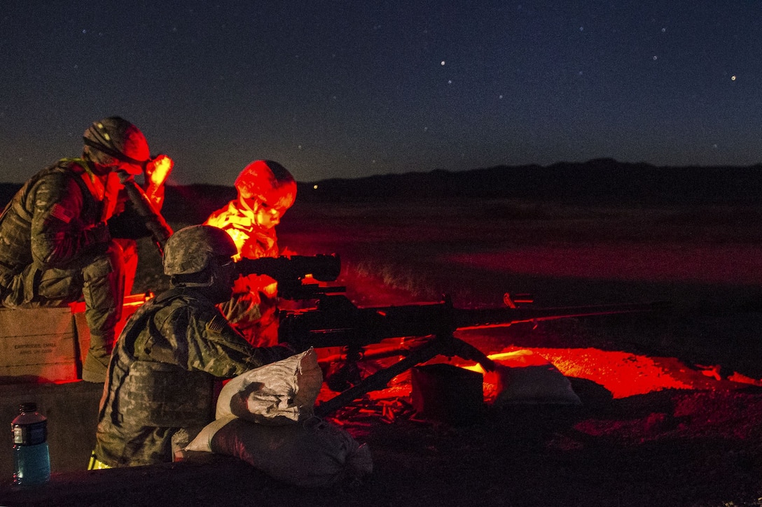 Soldiers illuminated by red light gather behind a machine gun outside at night.