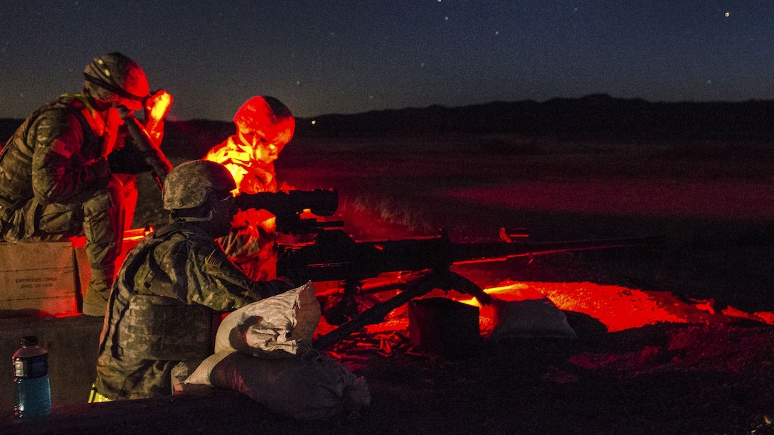 Soldiers illuminated by red light gather behind a machine gun outside at night.