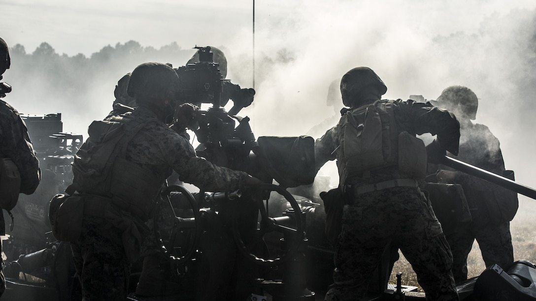 Smoke surrounds a howitzer, as a Marine looks through its sight and others and others work nearby.
