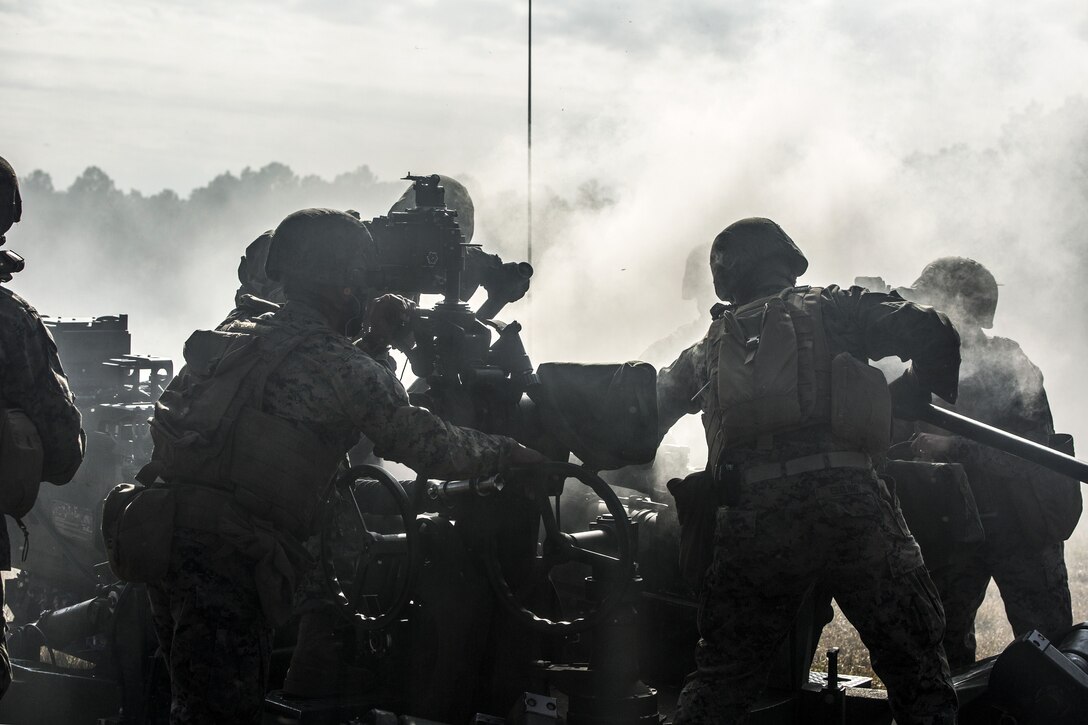 Smoke surrounds a howitzer, as a Marine looks through its sight and others and others work nearby.