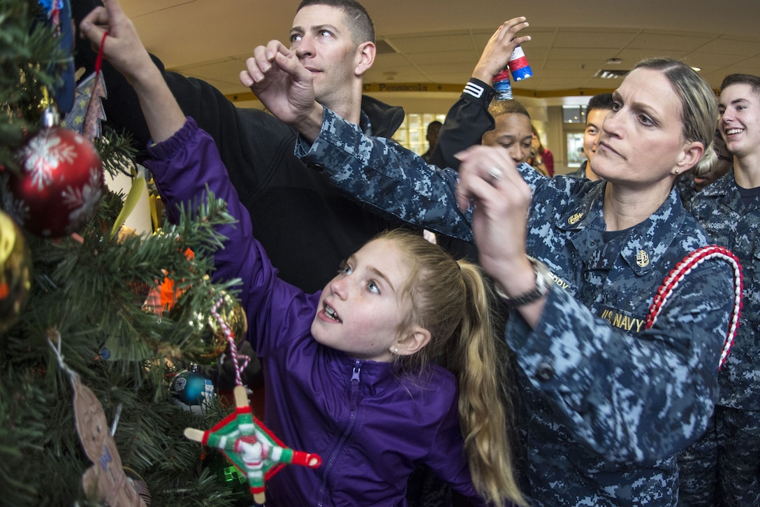 A girl reaches with an ornament toward a Christmas tree as a woman stands behind her with her arms extended.