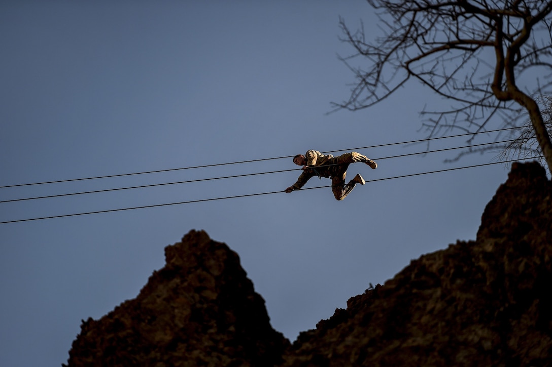 A soldier crawls across a high wire over a rocky landscape.