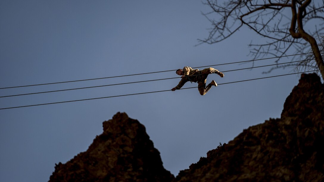 A soldier crawls across a high wire over a rocky landscape.