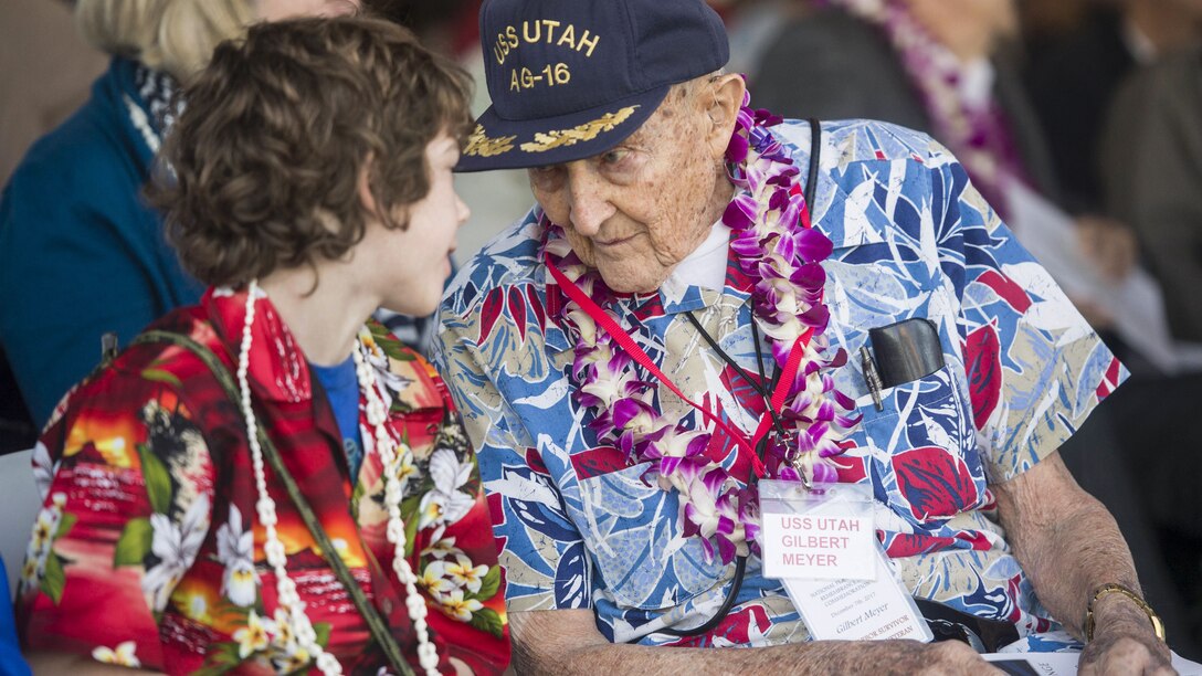 An elderly man wearing a Hawaiian print shirt and a lei, speaks to a boy who is similarly dressed.