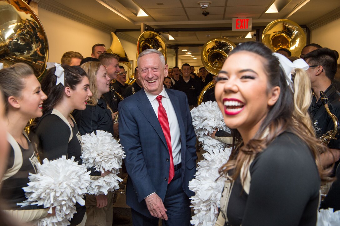 Defense Secretary James N. Mattis laughs while walking in a hallway crowded with musicians and cheerleaders.