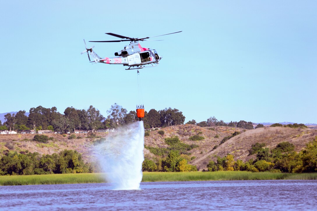 A UH-1Y Venom helicopter practices dropping water from a bucket firefighting system.