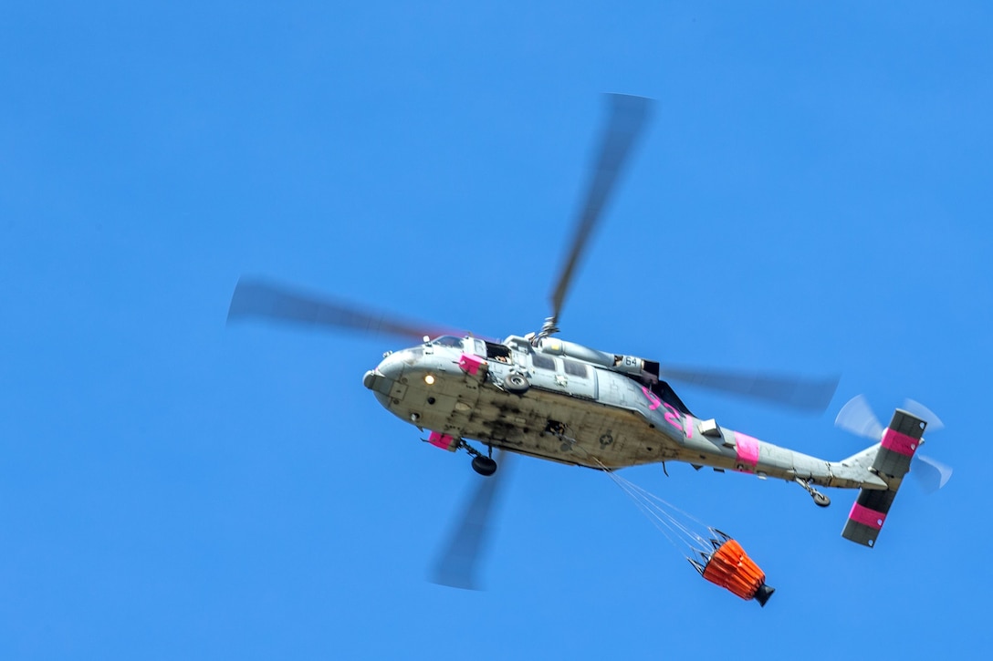 A helicopter flies against blue sky with a bucket attached to the bottom.