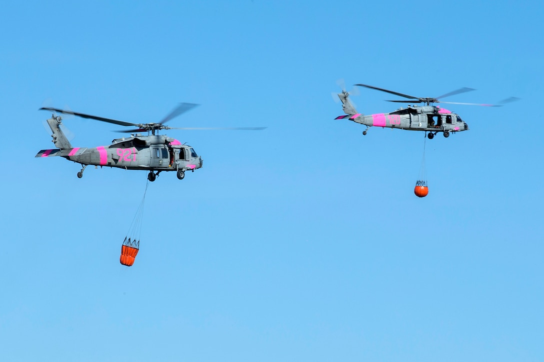 Two helicopters fly with buckets attached to them.
