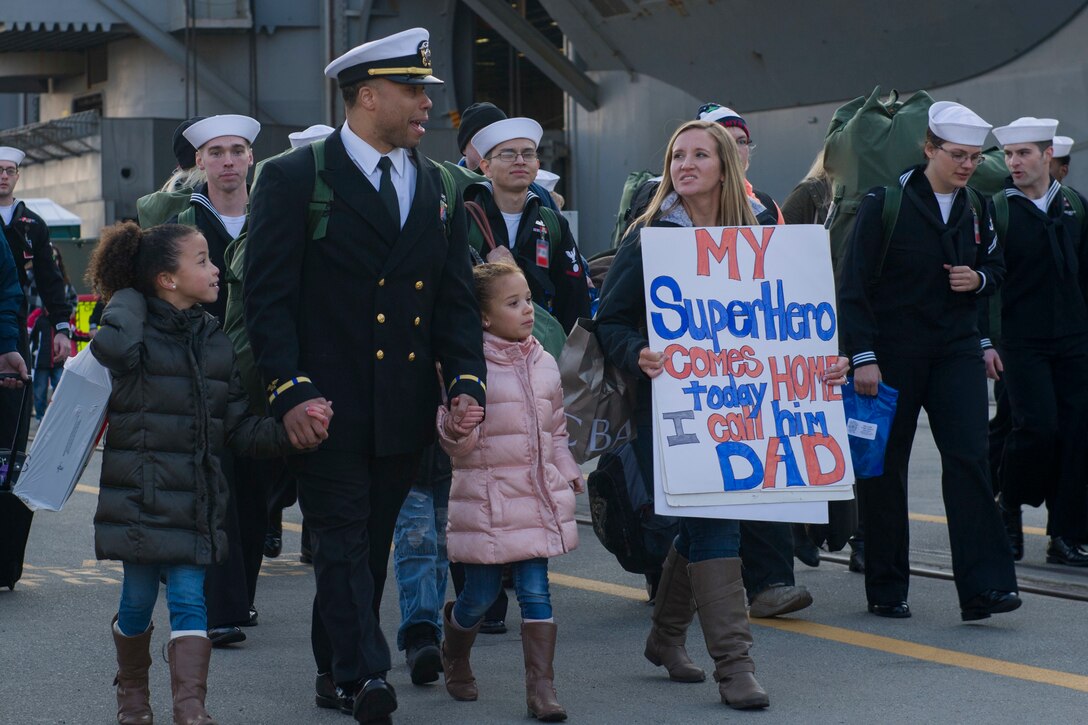 Sailors, children and families walk across the deck of a ship.