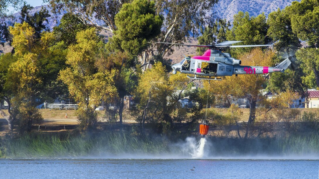 A helicopter fills an orange bucket with water from a lake.