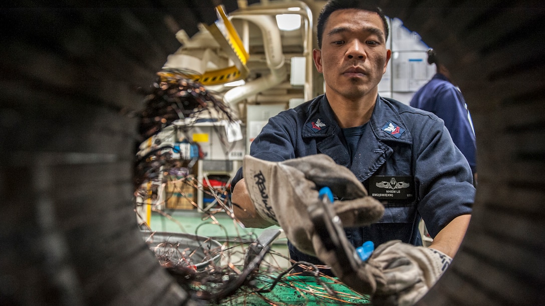 A sailor reaches with a gloved hand holding a tool into a circular opening of a motor.