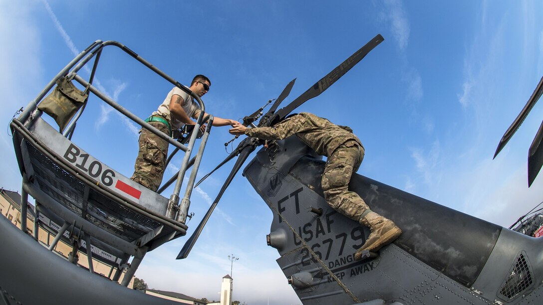 One airman straddles the tail of a helicopter and reaches toward another airman on a raised platform.