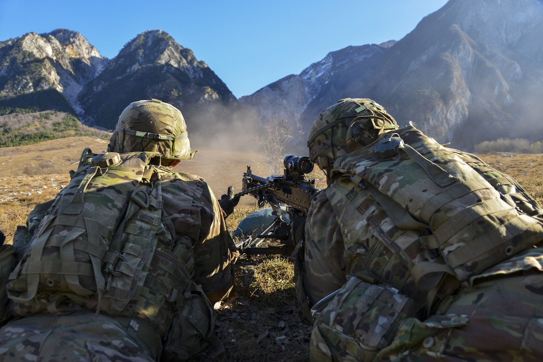 Two soldiers, visible from behind, fire a weapon in the direction of a mountain range in the distance.