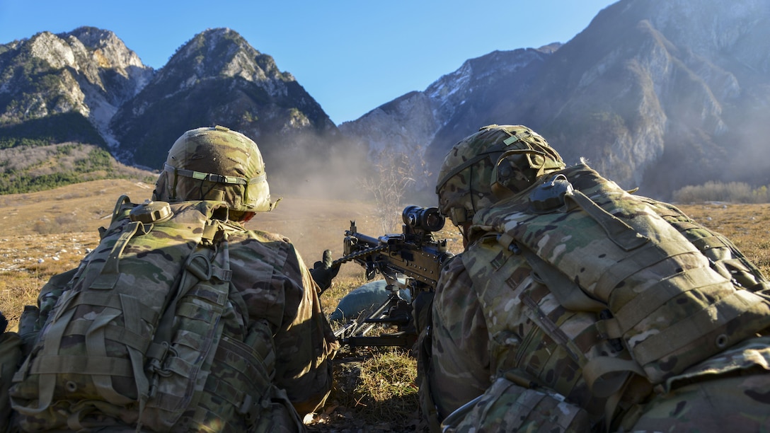 Two soldiers, visible from behind, fire a weapon in the direction of a mountain range in the distance.