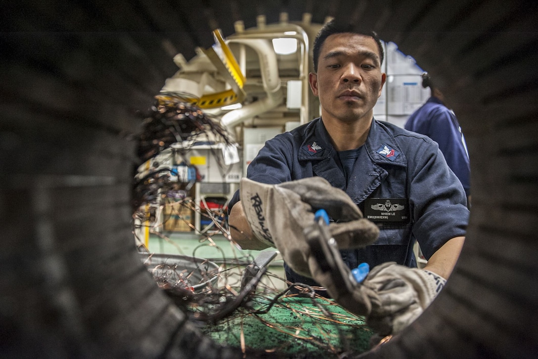 A sailor reaches with a gloved hand holding a tool into a circular opening of a motor.