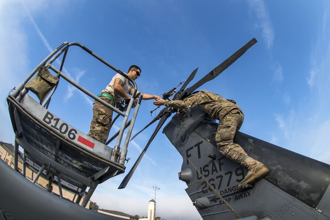 One airman straddles the tail of a helicopter and reaches toward another airman on a raised platform.