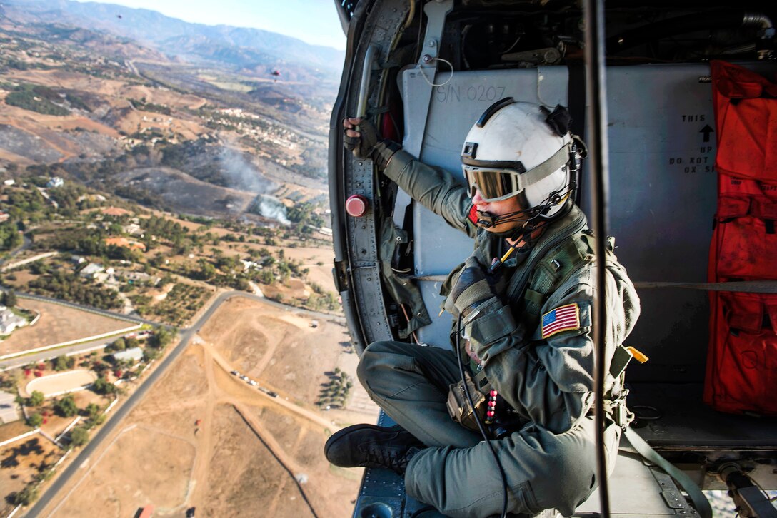 A service member sits on the edge of a helicopter looking for wildfires.