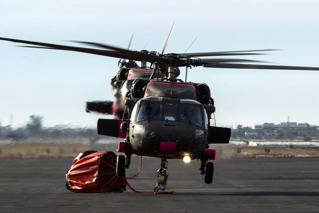 Two UH-60 Black Hawk helicopters land with attached bucket firefighting systems.