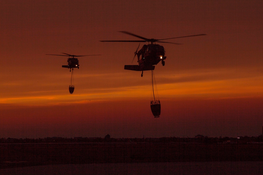 Two Army UH-60 Black Hawk helicopters land at sunset with bucket firefighting equipment