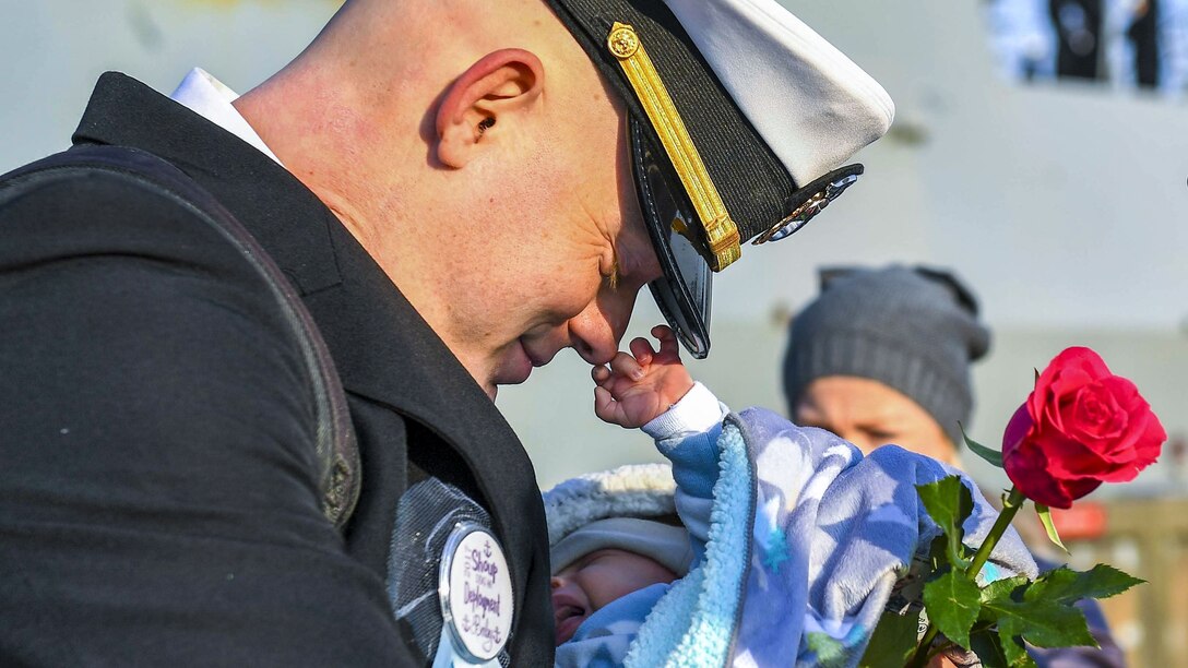 A smiling sailor bows his head with eyes closed as a baby he's holding touches his nose.