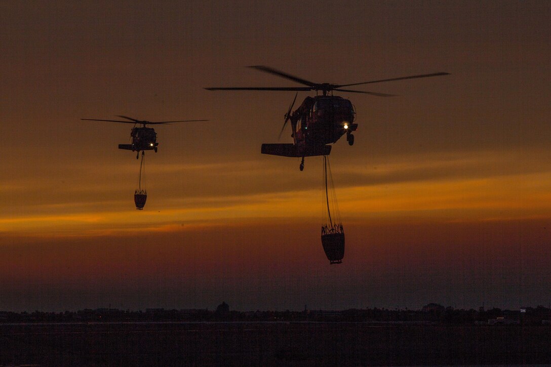 Two helicopters with attached water buckets fly against an orange sky.
