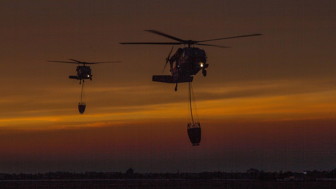 Two helicopters with attached water buckets fly against an orange sky.