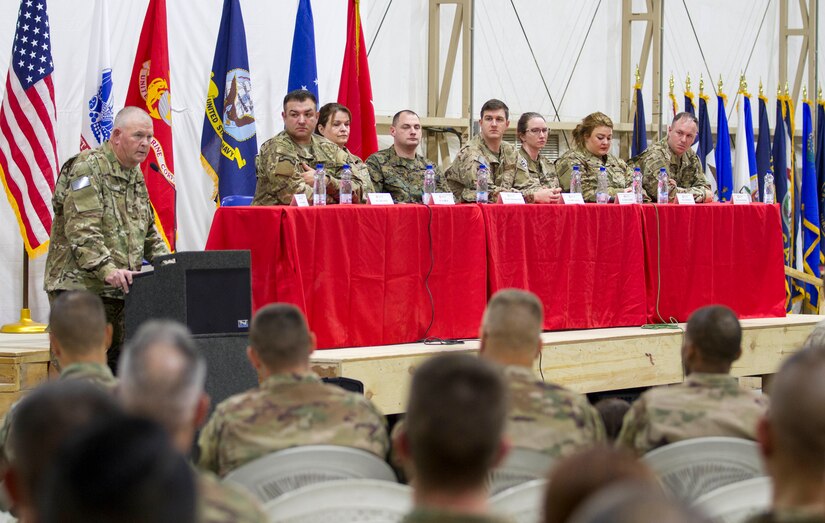 Soldiers sit on stage while another gives a speech.