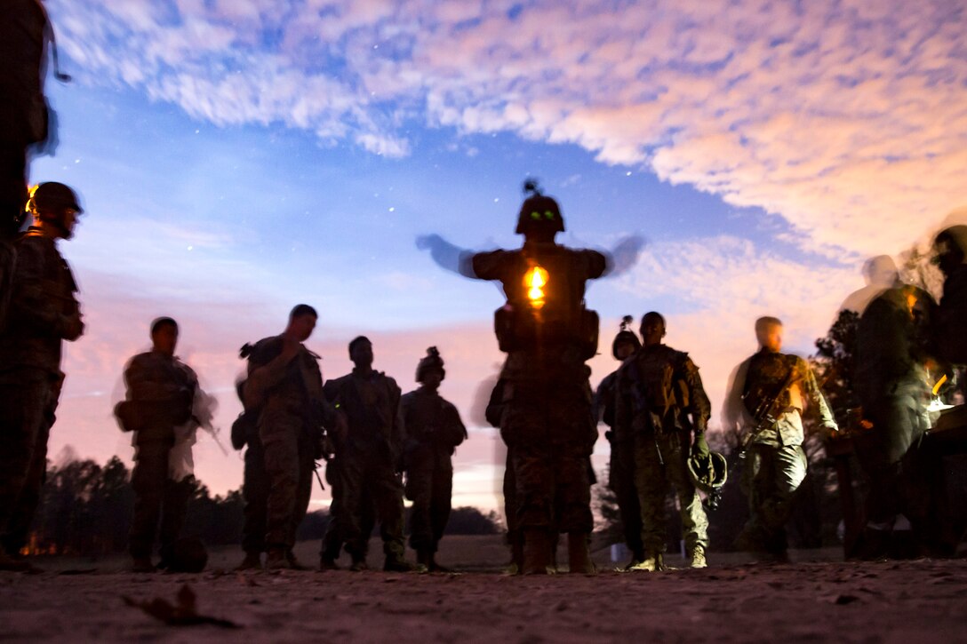 Marines, shown in silhouette, gather around a fellow Marine against a starry blue sky with pink clouds.