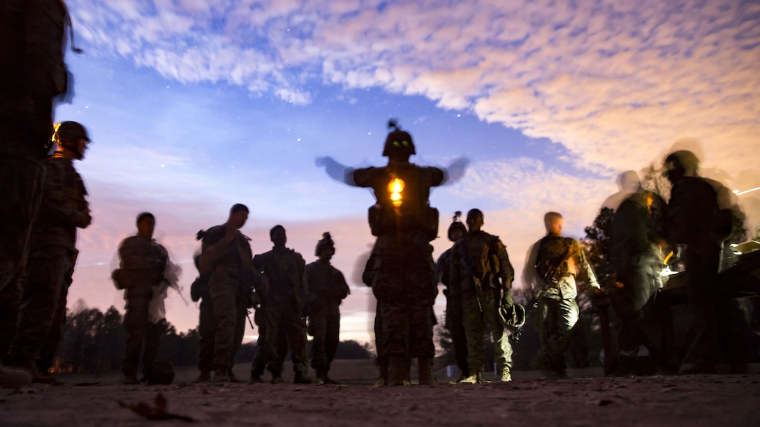 Marines, shown in silhouette, gather around a fellow Marine against a starry blue sky with pink clouds.