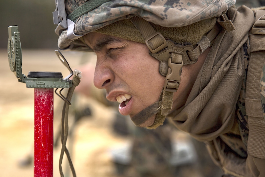 A Marine, shown in close-up, squints through an optical device mounted on a red pole.