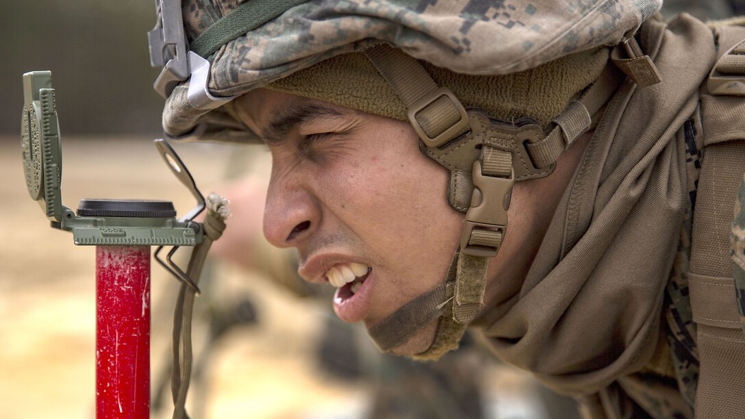 A Marine, shown in close-up, squints through an optical device mounted on a red pole.