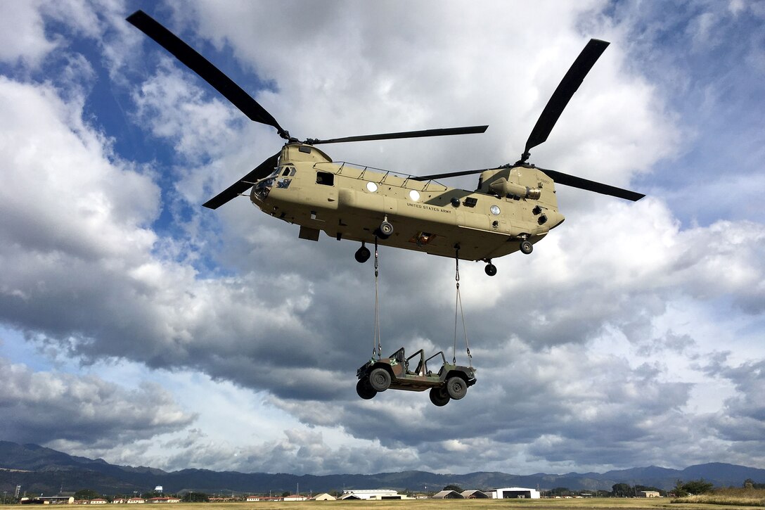 A military vehicle hangs by sling lines from a hovering Chinook helicopter.