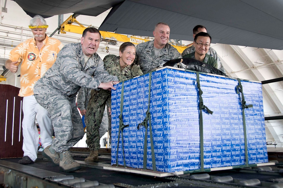 Service members push a pallet containing a big blue gift-wrapped box down a ramp.